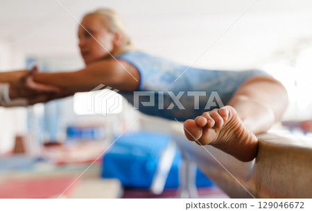 Woman gymnast performs complex acrobatic exercise on pommel horse with support of trainer in gym 129046672