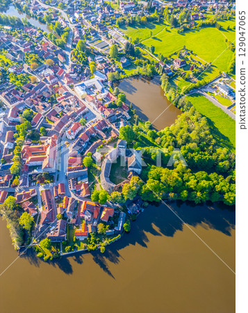 An aerial view captures the stunning castle of Rozmital pod Tremsinem surrounded by a quaint town, lush greenery, and a serene body of water during a clear day. An aerial view captures the stunning castle of Rozmital pod Tremsinem surrounded by a quaint town, lush greenery, and a serene body of water during a clear day. 129047065
