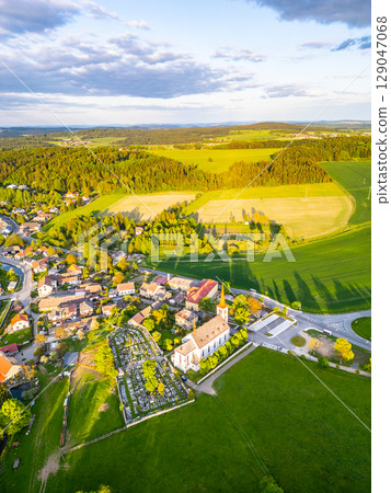 Bohutin Town is captured from above, showcasing its charming layout. Green fields stretch into the distance, complemented by a historic church and a cemetery. The sun casts long shadows. 129047068