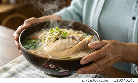 Samgyetang held in the hands of a Korean grandmother wearing traditional hanbok in a warmly lit home kitchen environment 129049143