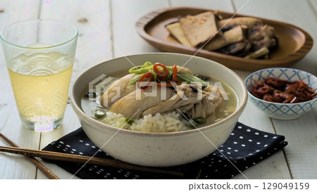 A bowl of korean samgyetang with side dishes and a glass of clear liquid 129049159