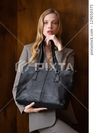 A stylish young woman, long blonde hair, poses in a grey suit, holding a black leather bag against a warm wooden background 129050375