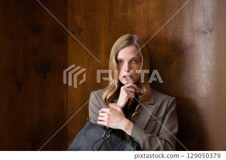 A stylish young woman, long blonde hair, poses in a grey suit, holding a black leather bag against a warm wooden background A stylish young woman, long blonde hair, poses in a grey suit, holding a black leather bag against a warm wooden background 129050379
