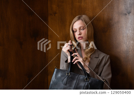 A stylish young woman, long blonde hair, poses in a grey suit, holding a black leather bag against a warm wooden background 129050400