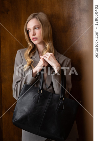 A stylish young woman, long blonde hair, poses in a grey suit, holding a black leather bag against a warm wooden background 129050402