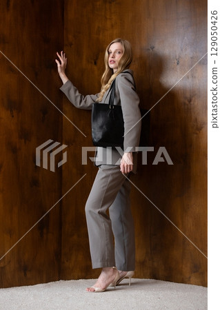 A stylish young woman, long blonde hair, poses in a grey suit, holding a black leather bag against a warm wooden background A stylish young woman, long blonde hair, poses in a grey suit, holding a black leather bag against a warm wooden background 129050426