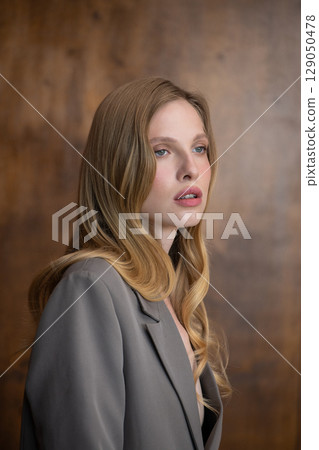 A beautiful young woman with long blonde wavy hair, wearing a stylish grey blazer, gazes thoughtfully to the side against a warm, blurred background 129050478