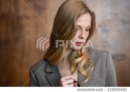 A beautiful young woman with long blonde wavy hair, wearing a stylish grey blazer, gazes thoughtfully to the side against a warm, blurred background A beautiful young woman with long blonde wavy hair, wearing a stylish grey blazer, gazes thoughtfully to the side against a warm, blurred background 129050482