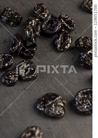 black sweet prunes made from small plums, piled on a board made of black stone dried plum fruits, close up 129051700