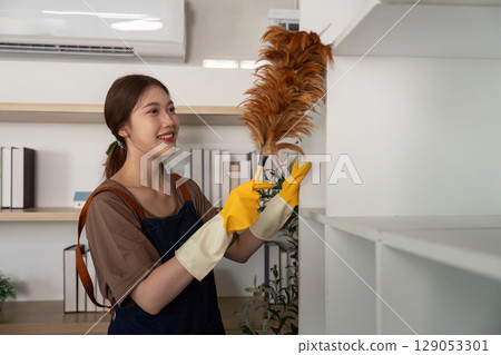 Home Cleaning. Woman dusting shelves with a feather duster and smiling. Home Cleaning. Woman dusting shelves with a feather duster and smiling. 129053301
