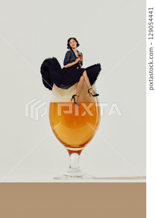 Woman in dark dress sitting playfully atop giant beer glass 129054441