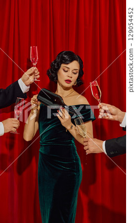 Woman in velvet dress pouring champagne from bottle into glass while surrounded by raised hands Woman in velvet dress pouring champagne from bottle into glass while surrounded by raised hands 129054452
