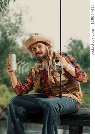 Bearded man in fishing outfit holding beer can and rod while smiling near water Bearded man in fishing outfit holding beer can and rod while smiling near water 129054453