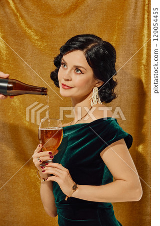 Smiling woman in velvet dress holding beer glass as bottle pours in front of golden curtain 129054455