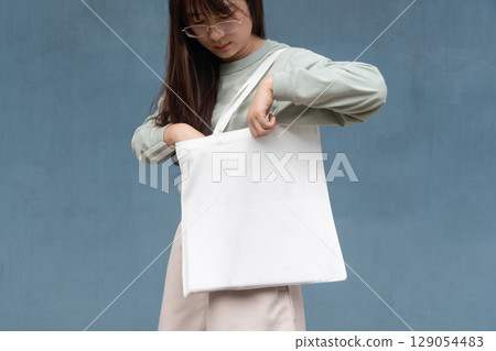 Blank white tote bag canvas fabric with handle mock up design. Close up of asian woman holding eco or reusable shopping bag against blue metal wall. No plastic bag and ecology concept. 129054483