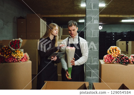 Young couple in warehouse preparing bouquets, surrounded by colorful floral arrangements and cardboard boxes, showcasing teamwork and creativity in floral design and arrangement process 129054584