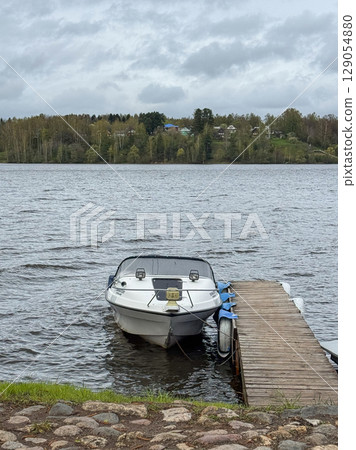 Boat moored at a wooden dock by a serene lake in a cloudy atmosphere Boat moored at a wooden dock by a serene lake in a cloudy atmosphere 129054880