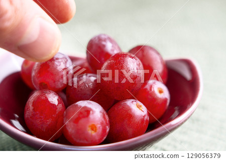 Ripe juicy red grapes and hand reaching for edible berries in ceramic bowl on green table Ripe juicy red grapes and hand reaching for edible berries in ceramic bowl on green table 129056379