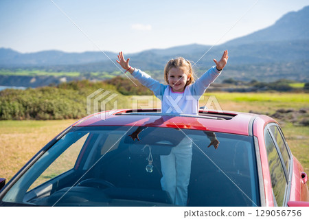 Little girl joyfully sticks out of car sunroof with raised hands against the backdrop of mountains in Northern Cyprus. The image symbolizes freedom, adventure, and the excitement of travel with child Little girl joyfully sticks out of car sunroof with raised hands against the backdrop of mountains in Northern Cyprus. The image symbolizes freedom, adventure, and the excitement of travel with child 129056756