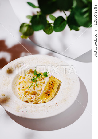 A sophisticated plate of creamy fettuccine pasta elegantly garnished with microgreens and a slice of bruleed cheese, presented on a white ceramic dish with parmesan dusting 129058288
