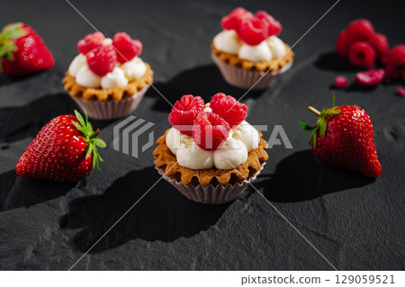 Delicious raspberry topped mini tarts with strawberries on a dark surface Delicious raspberry topped mini tarts with strawberries on a dark surface 129059521