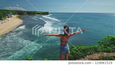 Woman standing on a cliff, arms spread wide, overlooking the serene waves and sandy shores of Dreamland Beach in Bali. Lush greenery and clear skies enhance the tranquil scene 129061529