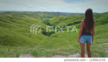 Young woman tourist is enjoying stunning view of rolling green hills and valleys in Sumba Island, Indonesia, feeling sense of freedom and adventure Young woman tourist is enjoying stunning view of rolling green hills and valleys in Sumba Island, Indonesia, feeling sense of freedom and adventure 129061530