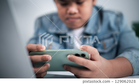 Young person holding green smartphone horizontally, focused on gaming or streaming, with a laptop in foreground, representing digital entertainment and modern youth technology. 129062028