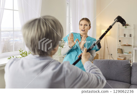 Angry elderly senior woman going to hit young female nurse with crutch. 129062236