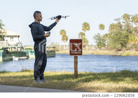 Operator is preparing to fly his quadcopter in national park no drone area. Man unlawfully using his UAV near restriction notice sign Operator is preparing to fly his quadcopter in national park no drone area. Man unlawfully using his UAV near restriction notice sign 129062856