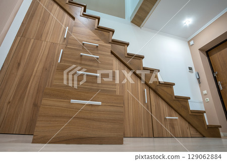 Modern architecture interior with luxury hallway with glossy wooden stairs in multi-storey house. Custom built pullout cabinets on glides in slots under stairs. Use of space for storage. Modern architecture interior with luxury hallway with glossy wooden stairs in multi-storey house. Custom built pullout cabinets on glides in slots under stairs. Use of space for storage. 129062884