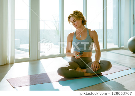 A woman is sitting on a yoga mat with her phone in her hand A woman is sitting on a yoga mat with her phone in her hand 129063707