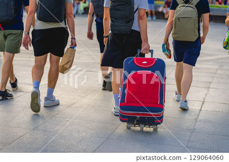Man with suitcase on wheels in city crowd. Man pulling red suitcase with wheels through busy pedestrian area. Concept of luggage in travel, active life style Man with suitcase on wheels in city crowd. Man pulling red suitcase with wheels through busy pedestrian area. Concept of luggage in travel, active life style 129064060