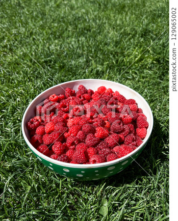 Bowl filled with fresh, ripe raspberries resting on vibrant green grass, showcasing the natural beauty and rich color of the berries in a sunny outdoor setting 129065002