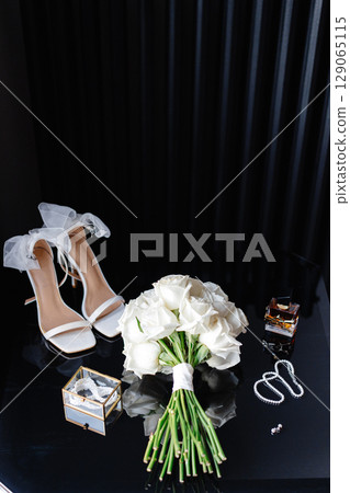 Elegant bridal accessories showcasing white roses bouquet, stylish heels, perfume, and jewelry on a dark glass table 129065115