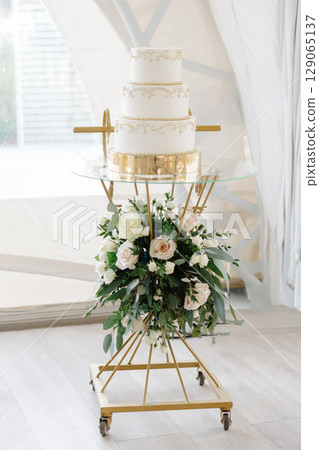Elegant white wedding cake decorated with gold standing on glass and gold stand with flowers Elegant white wedding cake decorated with gold standing on glass and gold stand with flowers 129065137