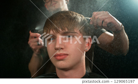 Young Man with Beard at Barber Shop. Barber is Preparing his Haircut. Dark Atmosphere, Black Background. Concept of Barber and a Client. 129065197