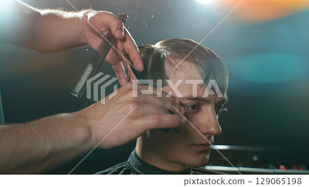 Young Man with Beard at Barber Shop. Barber is Preparing his Haircut. Dark Atmosphere, Black Background. Concept of Barber and a Client. 129065198