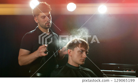 Young Man with Beard at Barber Shop. Barber is Preparing his Haircut. Dark Atmosphere, Black Background. Concept of Barber and a Client. 129065199