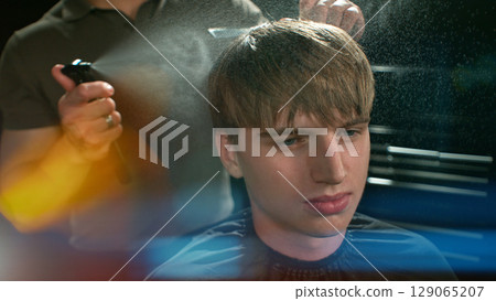 Young Man with Beard at Barber Shop. Barber is Preparing his Haircut. Dark Atmosphere, Black Background. Concept of Barber and a Client. 129065207