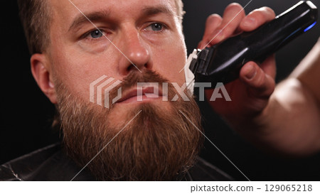 Young Man with Beard at Barber Shop. Barber is Preparing his Haircut. Dark Atmosphere, Black Background. Concept of Barber and a Client. 129065218