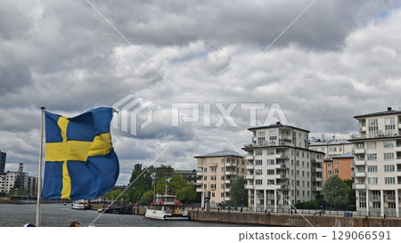 Swedish flag waving in the wind by the waterfront with boats and modern buildings in the background Swedish flag waving in the wind by the waterfront with boats and modern buildings in the background 129066591
