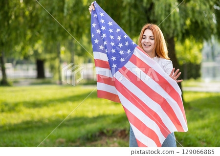 Happy young woman posing with USA national flag standing outdoors in summer park. Positive girl with United States banner outdoors. 129066885