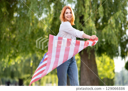 Happy young red haired woman posing with USA national flag standing outdoors in summer park. Positive girl celebrating United States independence day. Happy young red haired woman posing with USA national flag standing outdoors in summer park. Positive girl celebrating United States independence day. 129066886