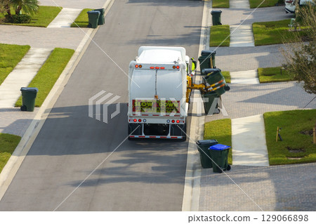 Garbage removal in residential area. Waste truck loading household rubbish while driving on suburban street Garbage removal in residential area. Waste truck loading household rubbish while driving on suburban street 129066898