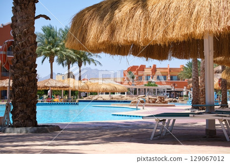 Empty deck chairs under straw shade umbrellas on swimming pool side in tropical resort. Summer vacations and getaway concept Empty deck chairs under straw shade umbrellas on swimming pool side in tropical resort. Summer vacations and getaway concept 129067012