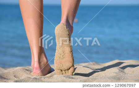 Close up of female feet walking barefoot on white grainy sand of golden beach on blue ocean water background Close up of female feet walking barefoot on white grainy sand of golden beach on blue ocean water background 129067097