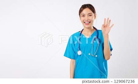 Happy smiling Asian woman nurse in blue uniform waving hello, a friendly welcome gesture from the young healthcare clinic nurse is shown, headshot isolated studio white background 129067236