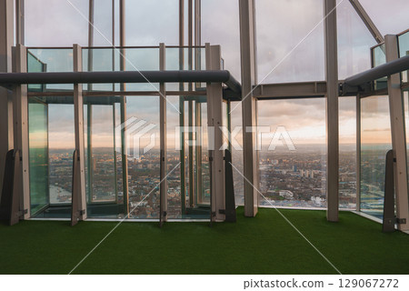The Shard's modern interior features large glass panels and green turf flooring, offering a panoramic view of London's cityscape under a cloudy sky. 129067272