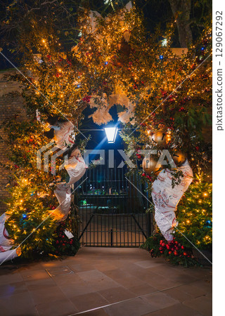 A decorated archway with golden bells, red berries, and greenery, illuminated by a street lamp. A wrought iron gate suggests a park entrance in London. A decorated archway with golden bells, red berries, and greenery, illuminated by a street lamp. A wrought iron gate suggests a park entrance in London. 129067292